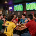 Group of friends enjoying live football matches while checking tỷ lệ kèo bóng đá at a lively sports bar.